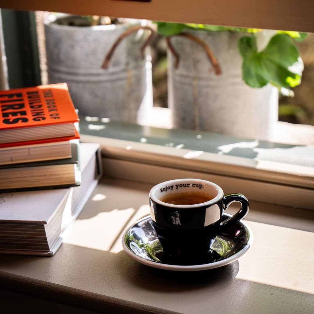 200 Degrees Coffee Shop coffee cup relaxing window books A cozy scene at 200 Degrees Coffee Shop featuring a small black coffee cup on a saucer, with the phrase "Enjoy your cup" printed inside. Nearby, a stack of colorful books rests on a sunlit windowsill, with potted plants in the background.