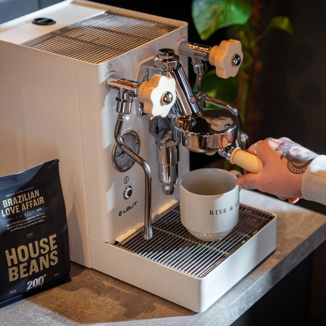 200 Degrees Coffee Shop espresso machine coffee beans barista brew A close-up of a coffee machine at 200 Degrees Coffee Shop, with a hand holding a portafilter above a white mug that reads "RISE & SHINE." A bag of coffee labeled "BRAZILIAN LOVE AFFAIR" is visible on the counter. The setting is warm and inviting, showcasing the coffee-making process.
