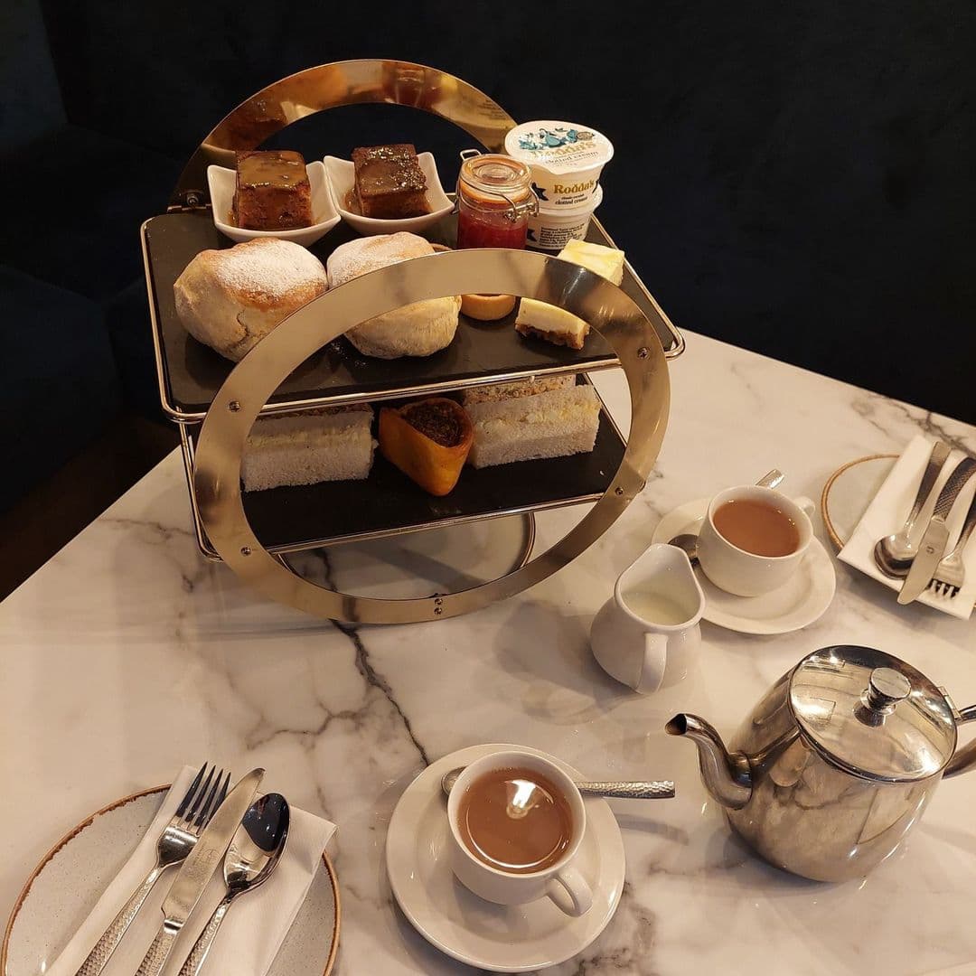 Convive Restaurant afternoon tea delights elegant setting gourmet treats A beautifully arranged afternoon tea set at Convive Restaurant, featuring a tiered stand with scones, pastries, and a selection of jams. Two cups of tea and a teapot are placed on a marble table, alongside polished cutlery and plates.