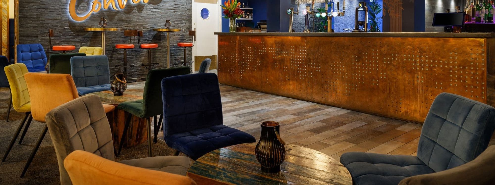 Alt text: Interior of Convive Restaurant featuring colorful chairs, a stylish bar area, and a warm, inviting atmosphere. The decor includes a textured wall and wooden tables, creating a modern yet cozy dining space.