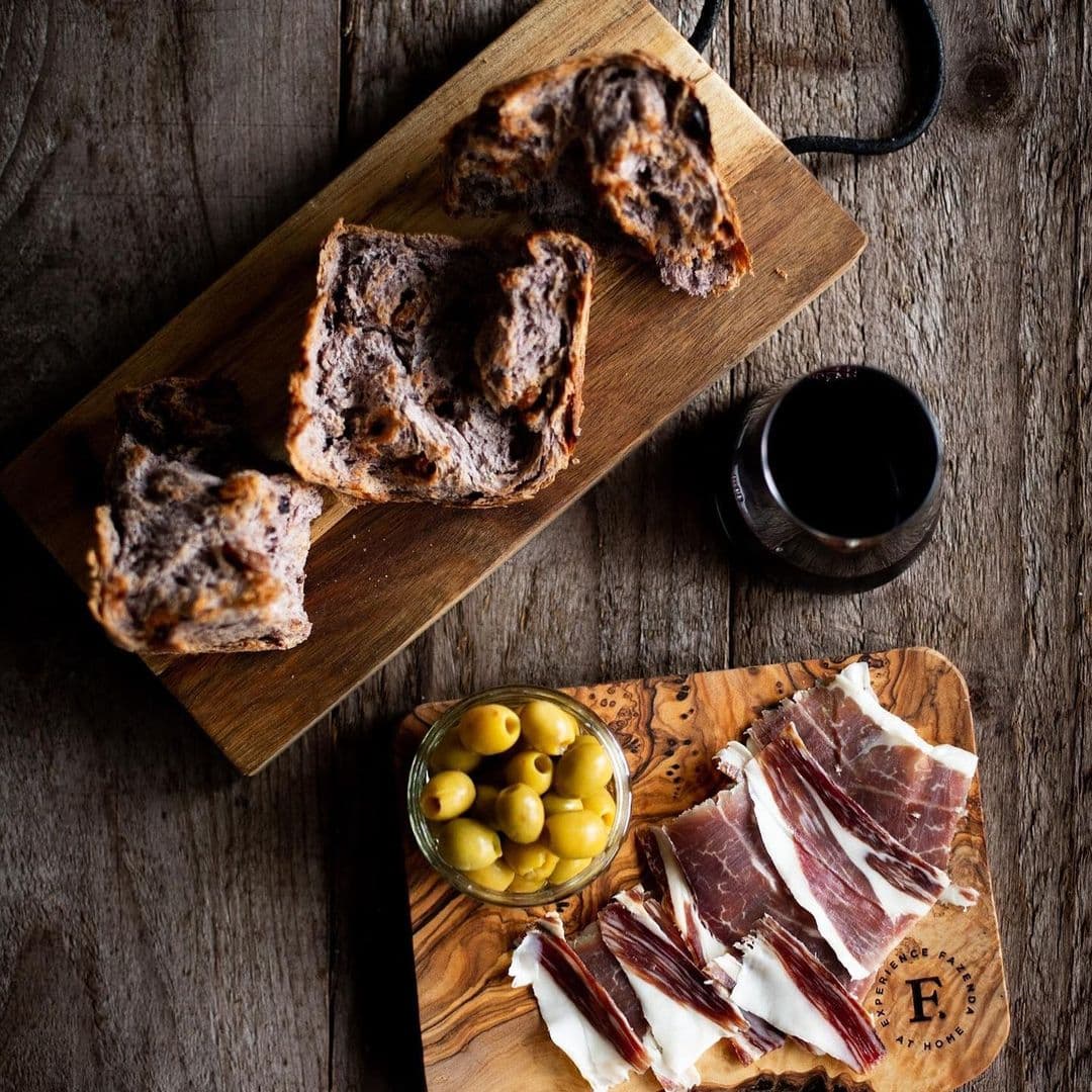 Fazenda Bar Grill artisanal bread charcuterie olives wine A wooden platter featuring slices of cured meat and a small jar of green olives, accompanied by a loaf of bread on a separate wooden board. A dark drink is placed nearby on a rustic wooden table. This image represents the culinary offerings at Fazenda Bar & Grill.