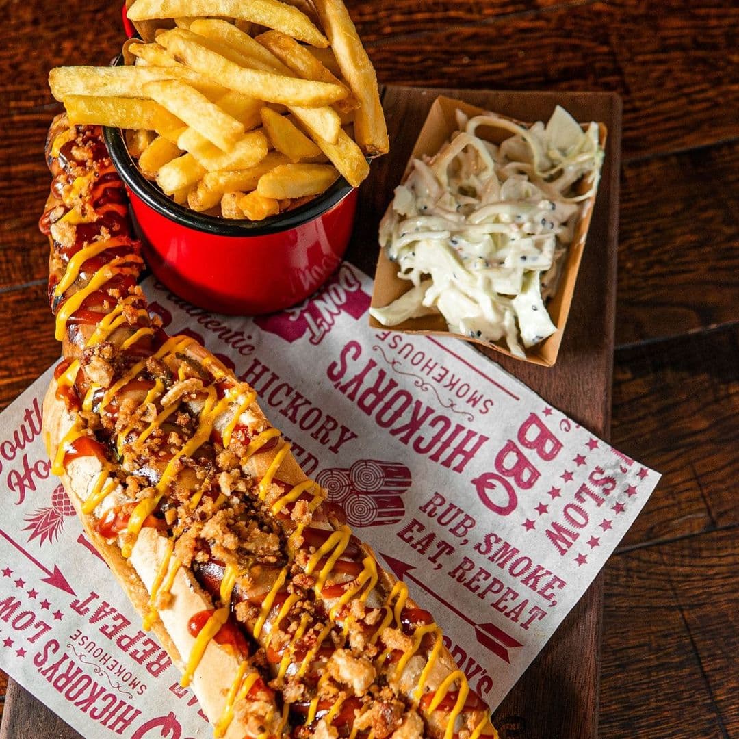 Fazenda Bar Grill bbq hotdog fries coleslaw delicious Delicious meal at Fazenda Bar & Grill featuring a large hot dog topped with mustard and ketchup, served with crispy French fries in a red cup and a side of coleslaw. The food is presented on decorative paper with barbecue-themed designs.