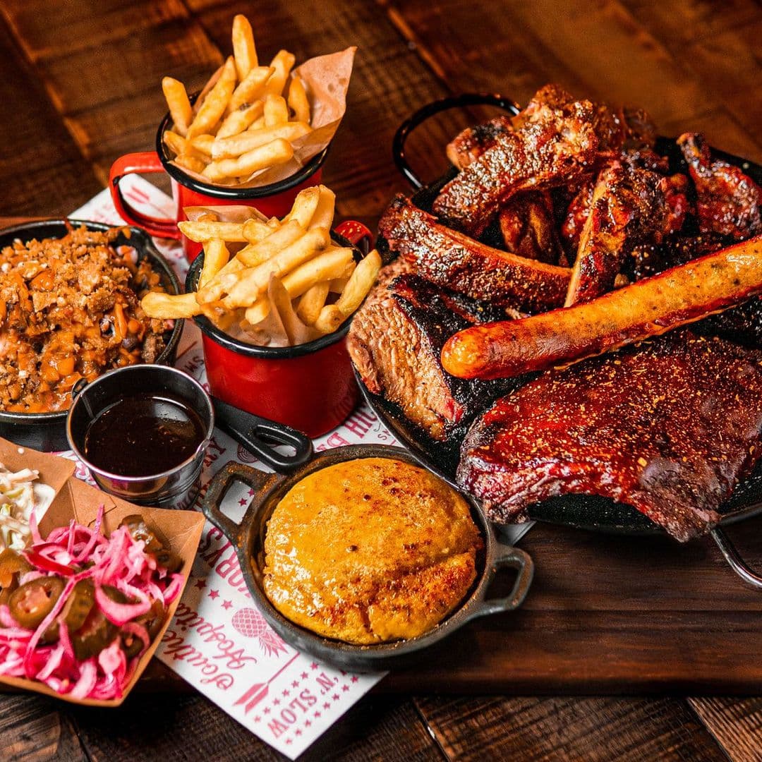 Fazenda Bar Grill bbq platter delicious food gastronomy A delicious spread of barbecue dishes at Fazenda Bar & Grill, featuring a variety of meats including ribs, sausage, and brisket. Accompanied by sides of crispy fries, baked beans, cornbread, pickled vegetables, and a small bowl of barbecue sauce. The meal is served on a wooden platter, showcasing the vibrant colors and textures of the food.
