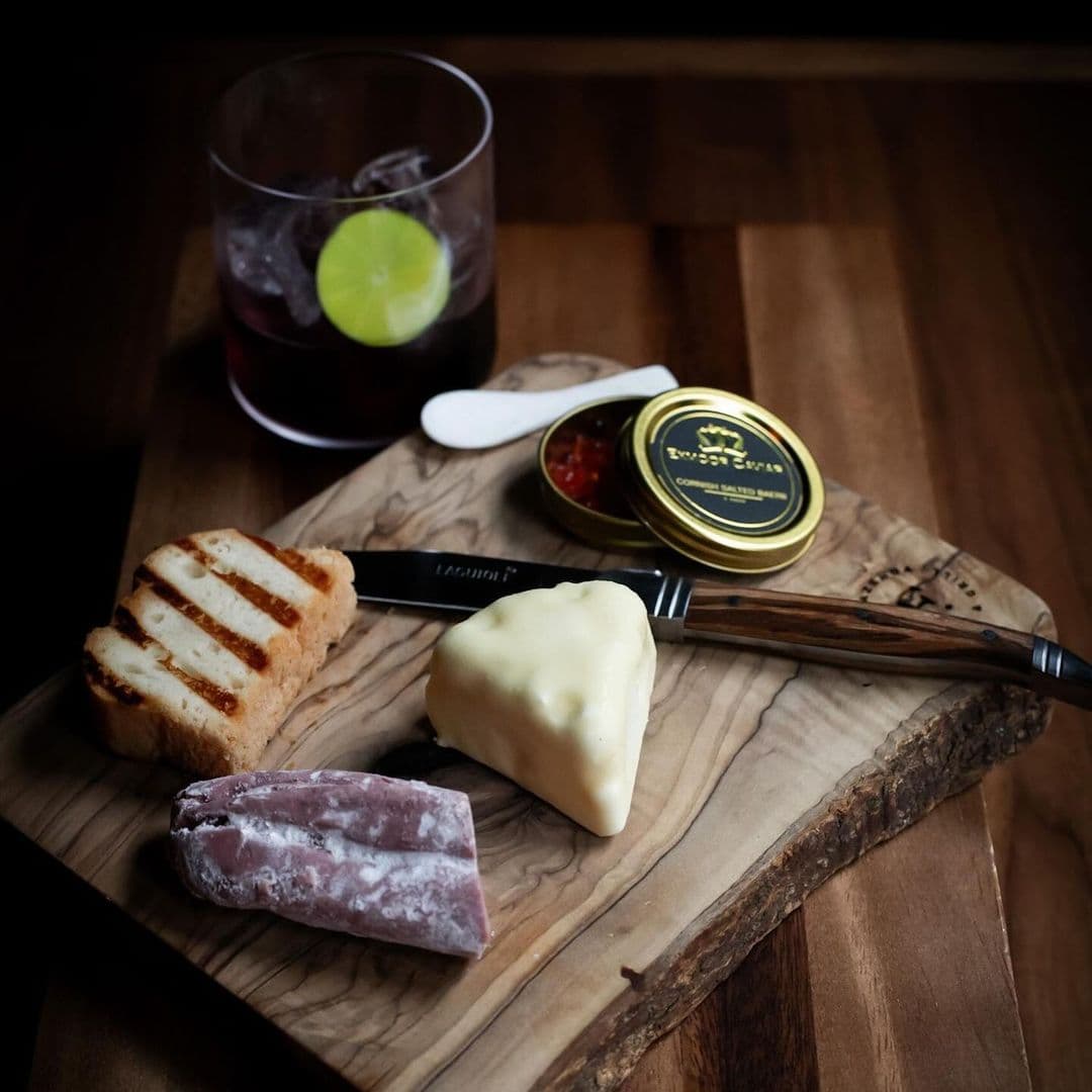 Fazenda Bar Grill cheese board drinks gourmet foods wooden platter A wooden serving board displays a selection of gourmet foods at Fazenda Bar & Grill. The arrangement includes grilled bread, a wedge of creamy cheese, a piece of cured meat, and a small tin of red condiment. A knife rests beside the food, and a glass of dark drink with a lime slice sits in the background.