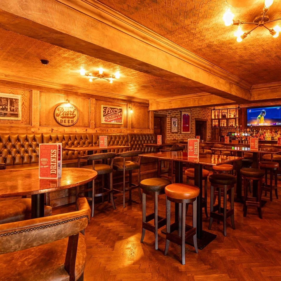 Fazenda Bar Grill cozy interior dining drinks ambiance Alt text: Interior of Fazenda Bar & Grill featuring wooden decor, cozy seating with leather accents, and tables set for dining. A television is visible in the background, and walls are adorned with vintage beer signs.