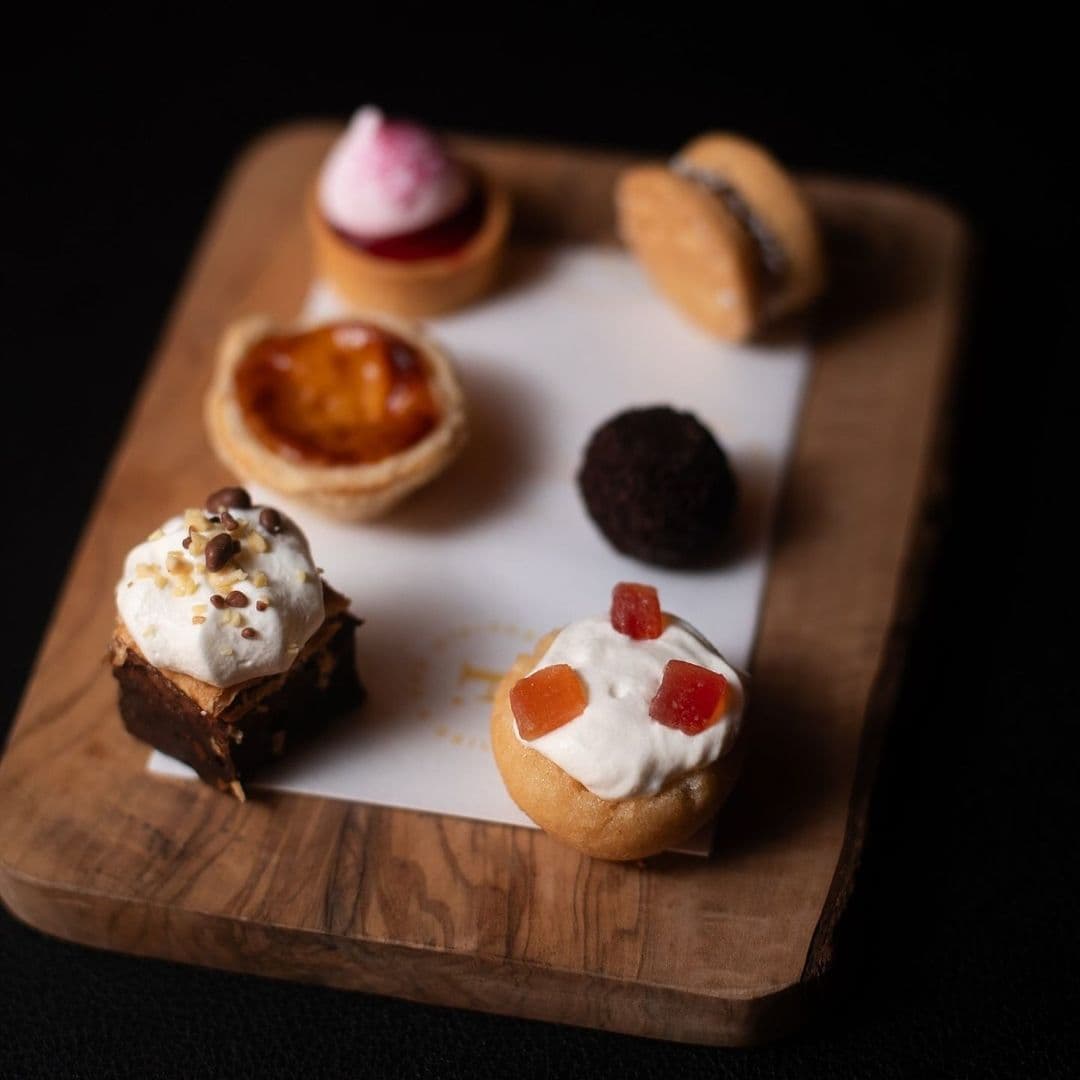 Fazenda Bar Grill desserts gourmet treats food platter A wooden platter displays an assortment of delightful desserts at Fazenda Bar & Grill. The selection includes a chocolate brownie topped with whipped cream and nuts, a small tart with caramel, a mini fruit tart, a chocolate truffle, and a sweet pastry with cream and colorful fruit pieces. The presentation highlights the variety and appeal of the desserts.