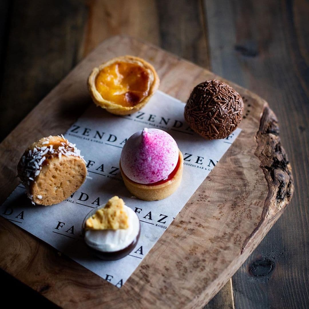 Fazenda Bar Grill desserts platter sweets gourmet treats A wooden platter displays a variety of colorful desserts, including a pink ice cream cone, a yellow custard tart, a chocolate truffle, and a coconut-covered treat. The desserts are arranged on a piece of paper featuring the name "Fazenda" prominently. This image showcases the delightful sweets available at Fazenda Bar & Grill.