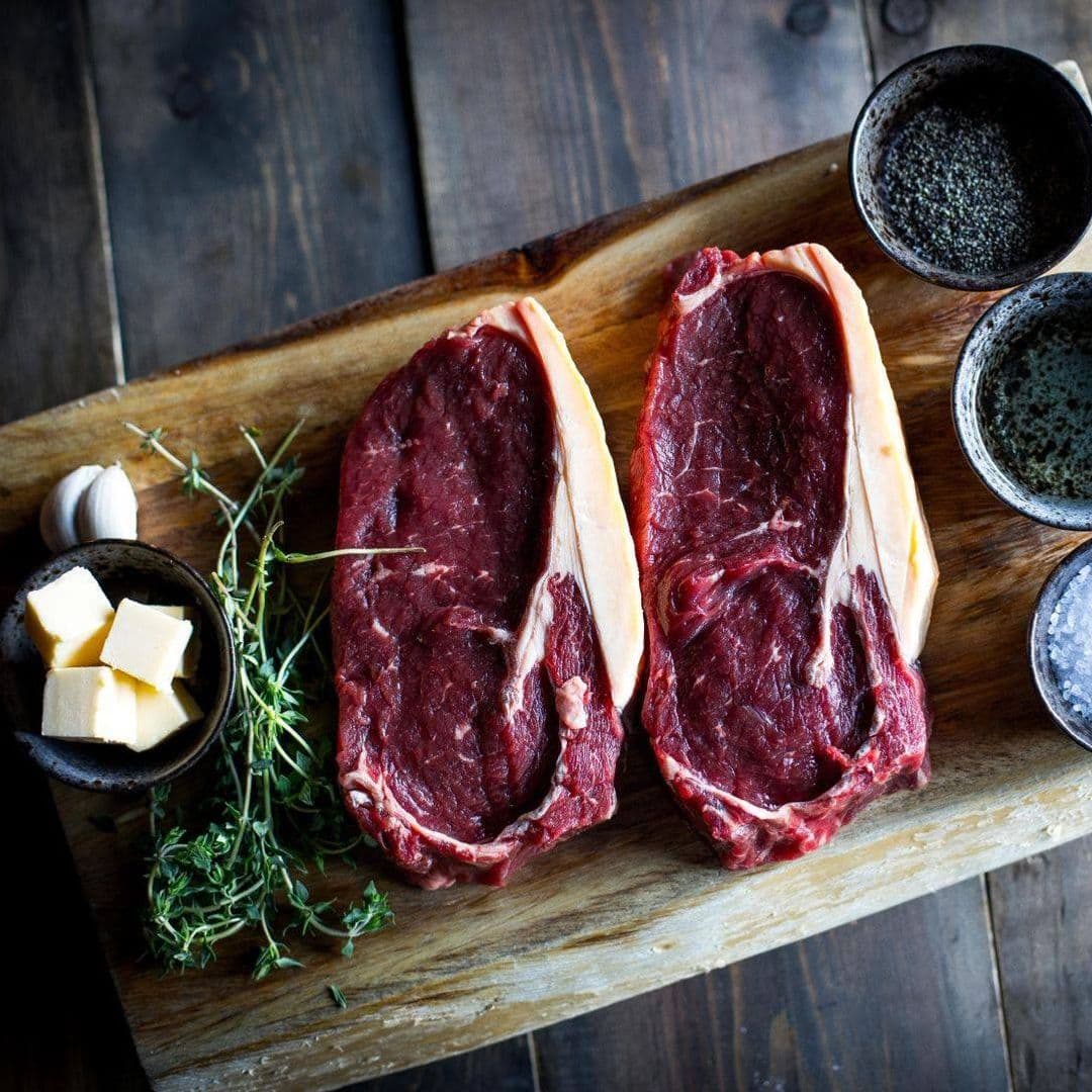 Fazenda Bar Grill gourmet steak fresh ingredients cooking preparation Two raw ribeye steaks are displayed on a wooden cutting board, accompanied by fresh thyme, garlic, and a small bowl of butter. Nearby, there are three bowls containing different types of seasoning, including salt and pepper. This image showcases the fresh ingredients available at Fazenda Bar & Grill.