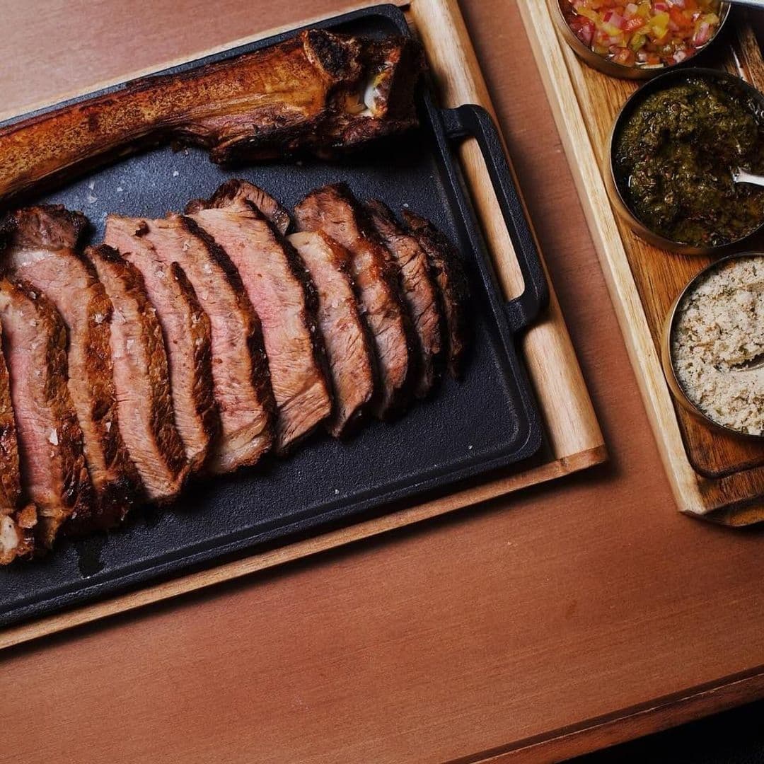 Fazenda Bar Grill grilled beef delicious meal sizzling platter Sliced grilled steak served on a black platter, accompanied by a bone and a wooden tray with bowls of salsa, green sauce, and seasoning at Fazenda Bar & Grill.
