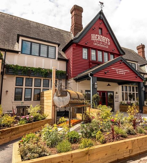 Fazenda Bar Grill hickorys smokehouse exterior garden dining A charming exterior view of Fazenda Bar & Grill, featuring a red and white building with large windows and a welcoming entrance. In the foreground, there are wooden planters filled with colorful flowers and greenery, adding to the inviting atmosphere. A smoker is visible outside, hinting at the delicious food offerings.