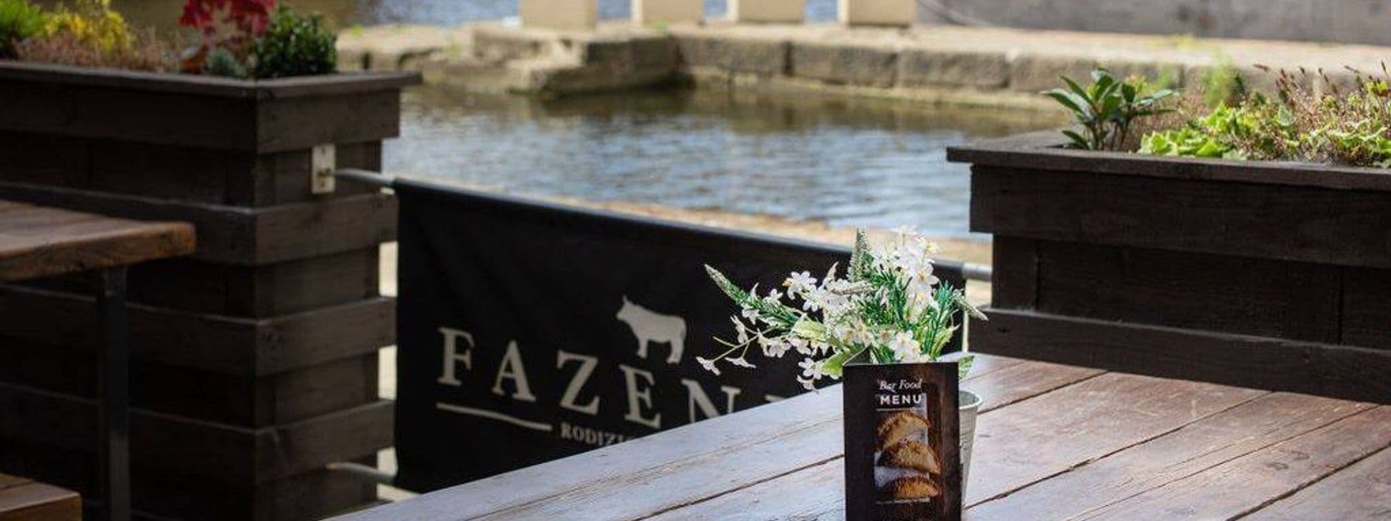 A table set for dining at Fazenda Bar & Grill, featuring a small vase with flowers and a menu. In the background, a calm water view and a black banner displaying the restaurant's name.