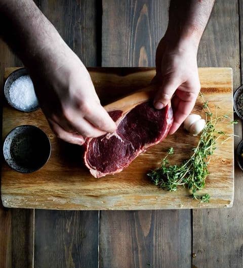 Fazenda Bar Grill preparing steak fresh ingredients cooking A person preparing a fresh cut of meat on a wooden cutting board, surrounded by bowls of salt and spices, along with herbs and an egg. This image represents the culinary experience at Fazenda Bar & Grill.