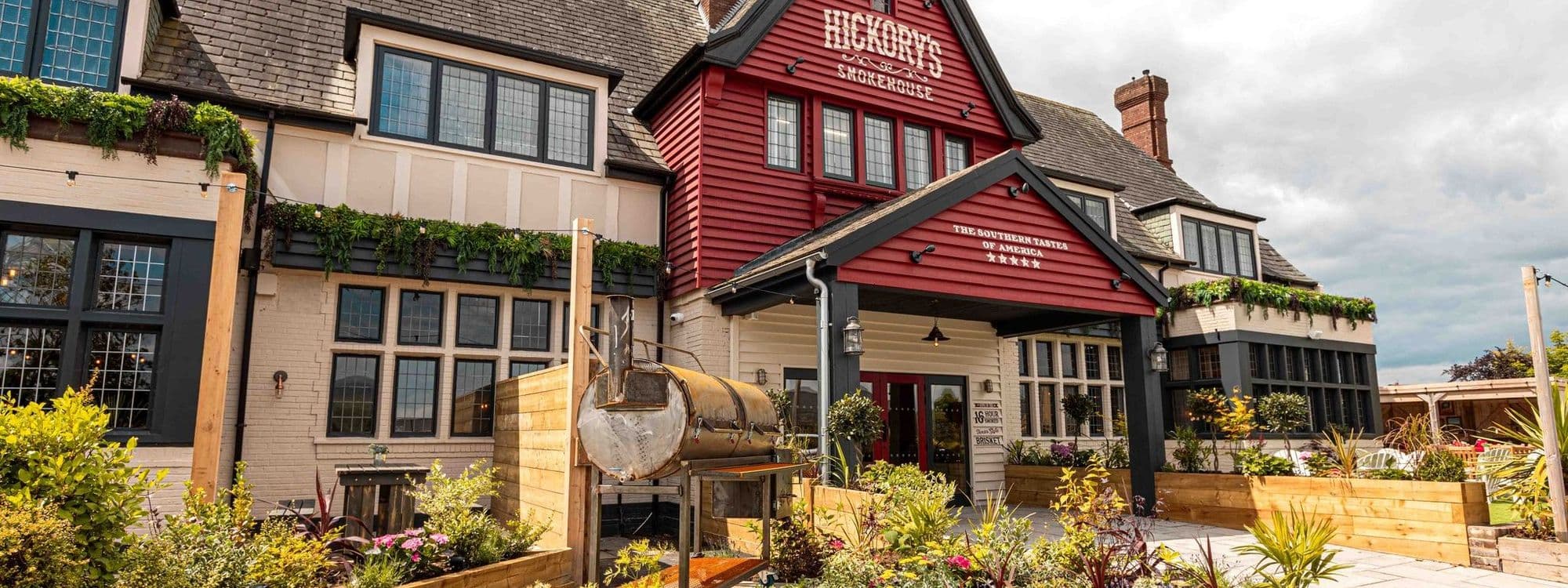Alt text: The exterior of Fazenda Bar & Grill features a charming building with a red and white facade, large windows, and decorative plants. The entrance is welcoming, with outdoor seating and lush greenery surrounding the area.