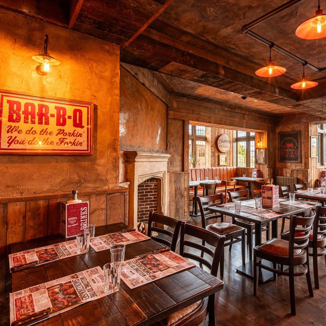 Fazenda Bar Grill rustic interior dining cozy ambiance barbecue Interior of Fazenda Bar & Grill featuring wooden tables and chairs, warm lighting, and a cozy atmosphere. The walls are decorated with rustic elements and a BAR-B-Q sign. Tables are set with menus and glasses, creating an inviting dining space.