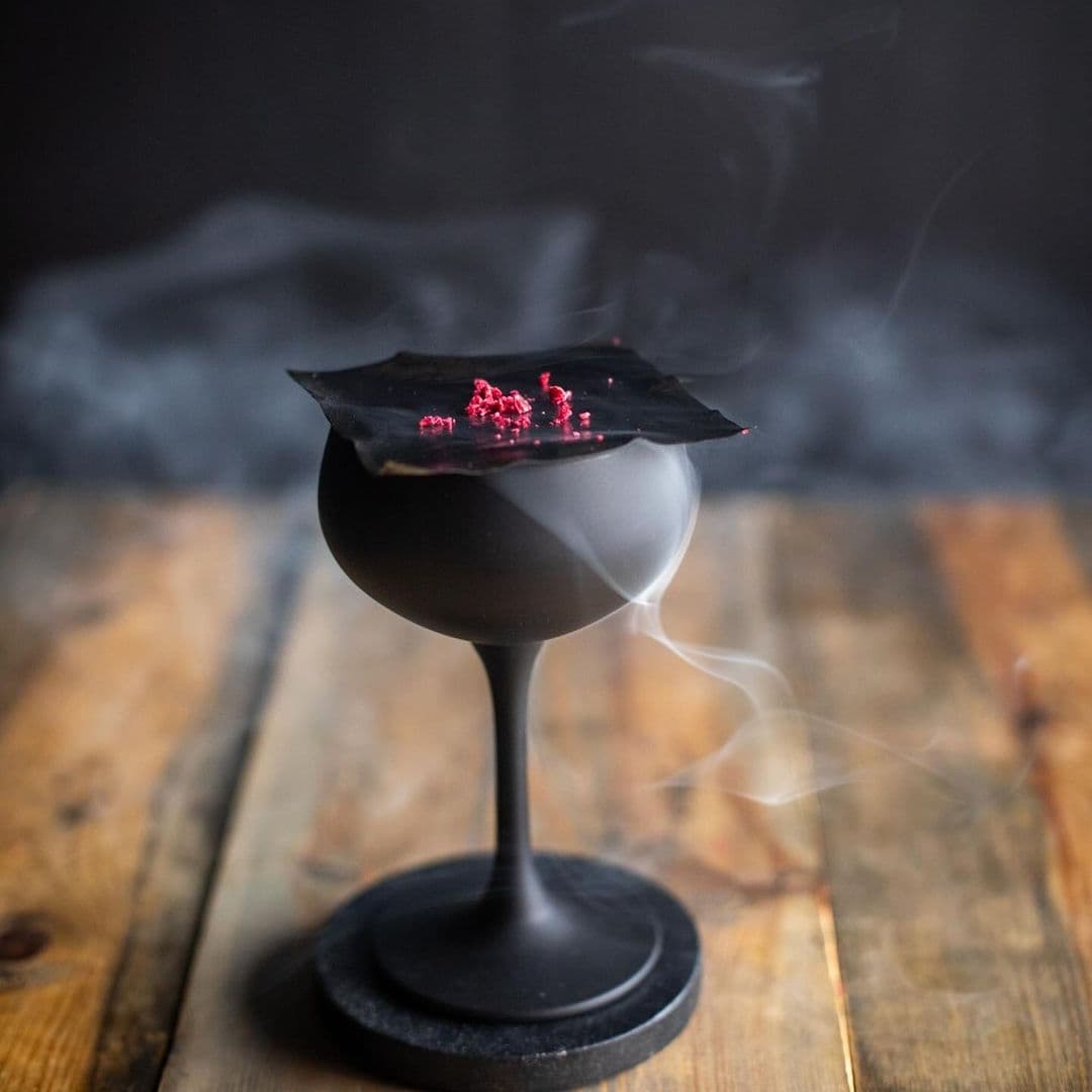 Fazenda Bar Grill smoky cocktail elegant presentation gourmet experience A stylish black cocktail glass topped with a dark sheet and vibrant red garnish, emitting wisps of smoke. The glass is set on a rustic wooden table, creating an inviting atmosphere at Fazenda Bar & Grill.