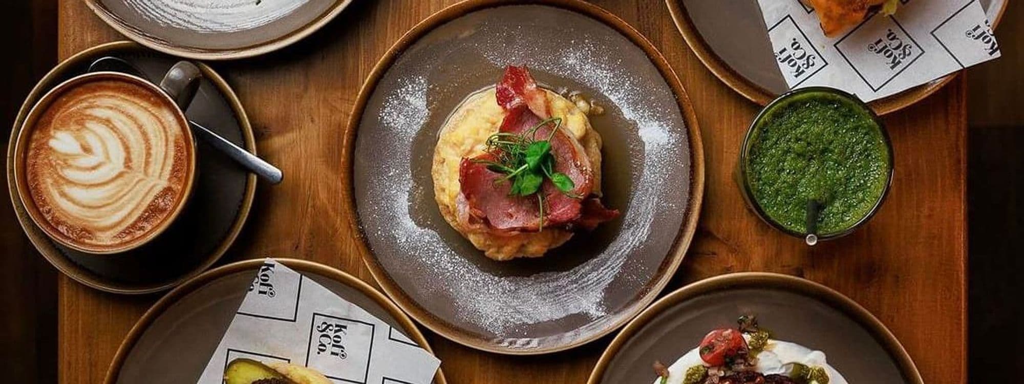 Delicious food spread at Kofi&Co – Roundhay, featuring a latte with latte art, a savory dish topped with ham and herbs, and various sides including a green sauce and a pastry. The setting is warm and inviting, perfect for a casual dining experience.