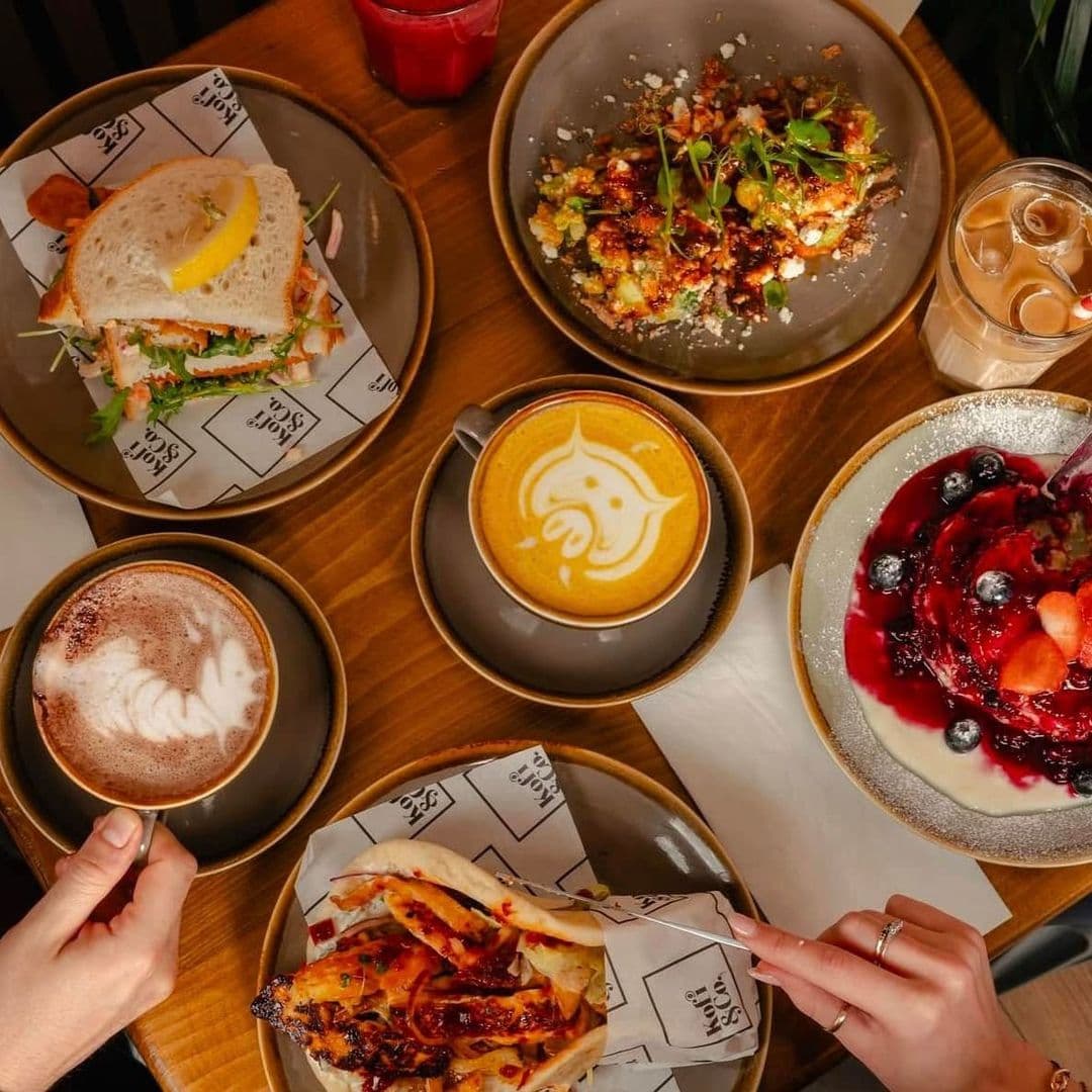 Kofi Co Roundhay delicious food coffee dessert brunch A beautifully arranged table at Kofi&Co – Roundhay featuring a variety of delicious food and drinks. The spread includes a sandwich with fresh ingredients, a colorful salad, a vibrant dessert topped with berries, and two cups of coffee with intricate latte art. A hand reaches for a dish, adding a personal touch to the inviting scene.