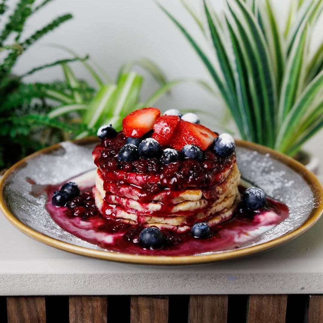 Kofi Co Roundhay pancakes berries syrup breakfast dessert Delicious stack of pancakes topped with mixed berry sauce, fresh strawberries, and blueberries, served on a decorative plate with a sprinkle of powdered sugar. Lush green plants in the background enhance the inviting atmosphere at Kofi&Co – Roundhay.