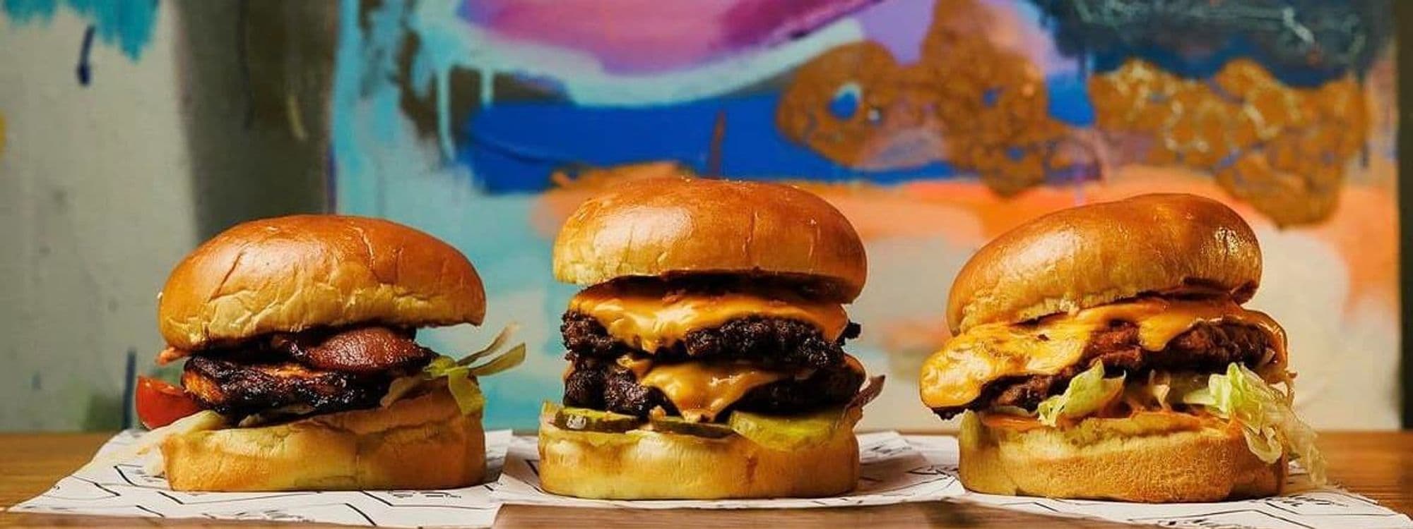 Three delicious burgers are displayed in front of a colorful background at Kofi&Co – Wetherby. Each burger features a toasted bun, melted cheese, and various toppings, showcasing a tempting selection for food lovers.