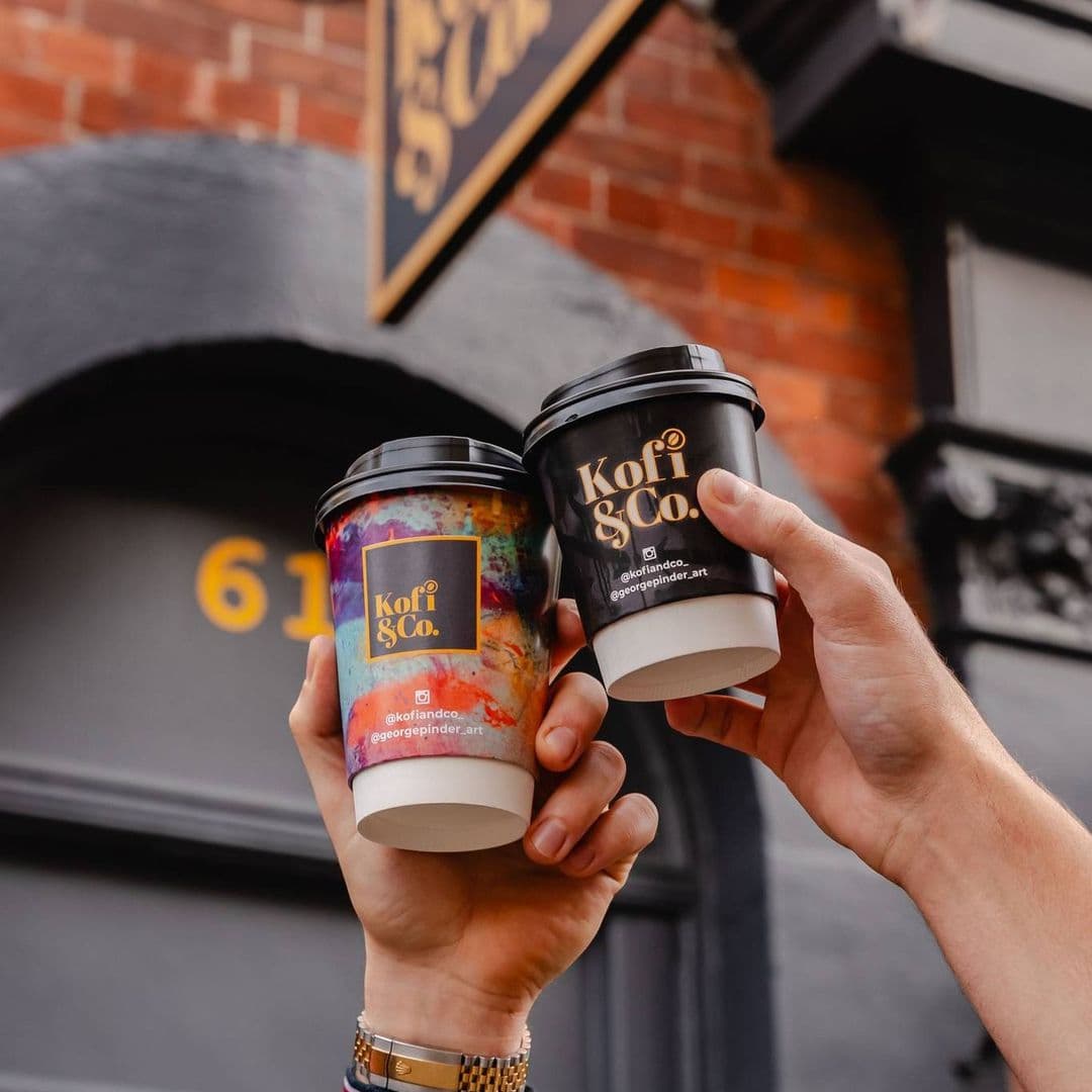 Kofi Co Wetherby coffee cups hand holding vibrant urban Two hands hold coffee cups from Kofi&Co, one with a colorful design and the other in a sleek black. The background features a brick wall with a sign for Kofi&Co – Wetherby.