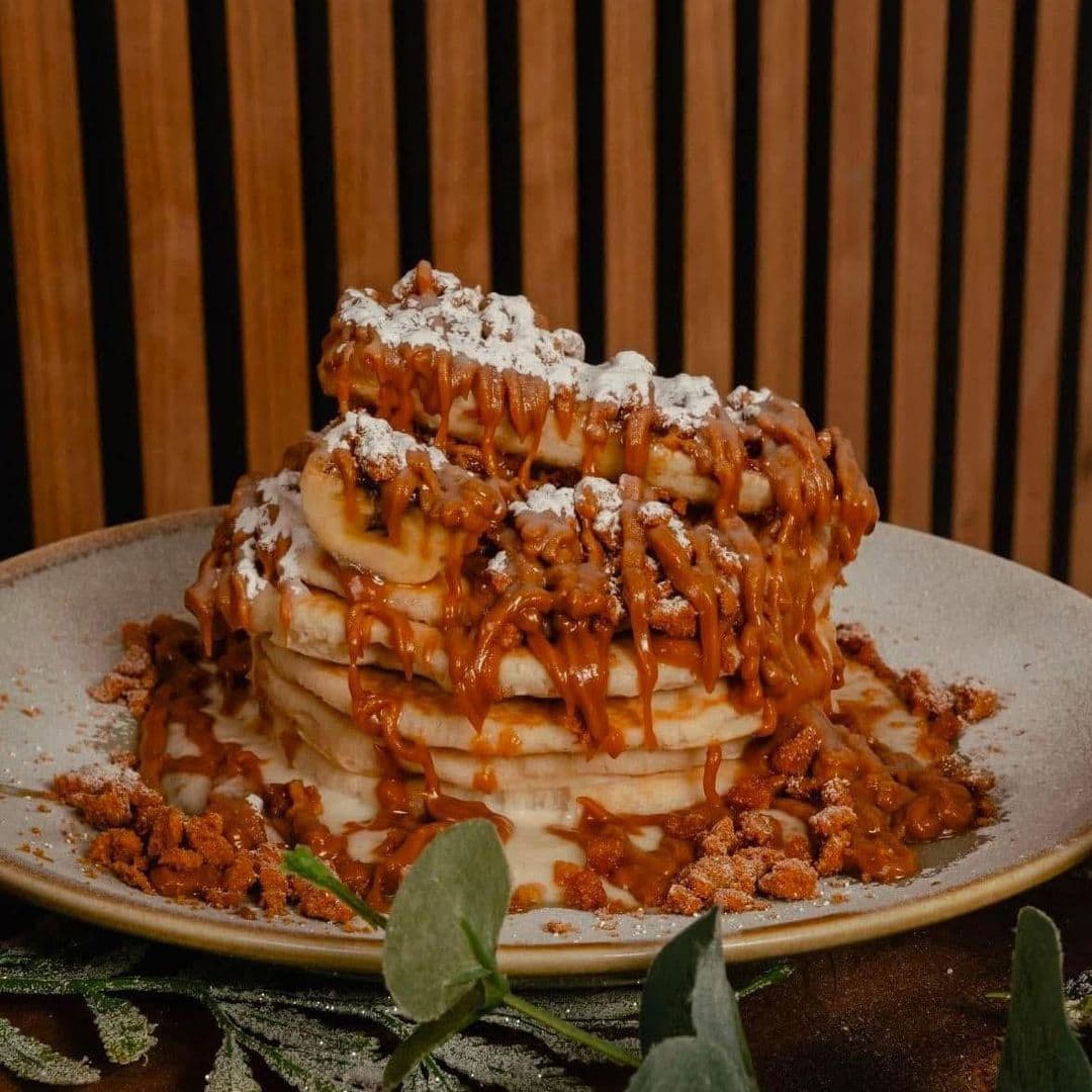 Kofi Co Wetherby pancakes delicious dessert brunch food Delicious stack of pancakes topped with caramel sauce, banana slices, and a dusting of powdered sugar, served on a plate with a sprinkle of crushed cookies. Kofi&Co – Wetherby.
