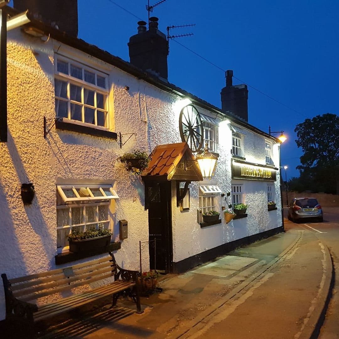 New Inn Barwick in Elmet cozy pub nighttime exterior warm lighting **Alt Text:** Exterior view of the New Inn – Barwick in Elmet, featuring a charming white building with warm lights, a wooden bench, and flower pots. The scene is set during twilight, highlighting the inviting atmosphere of the pub.