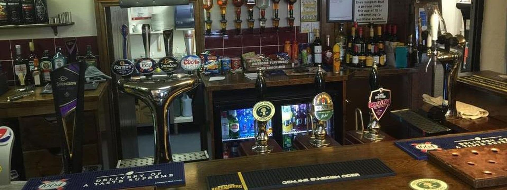 Alt text: Interior view of the bar at New Inn – Barwick in Elmet, featuring a variety of beer taps and a selection of spirits on the shelves. The wooden bar top is well-maintained, showcasing drinks and bar accessories.