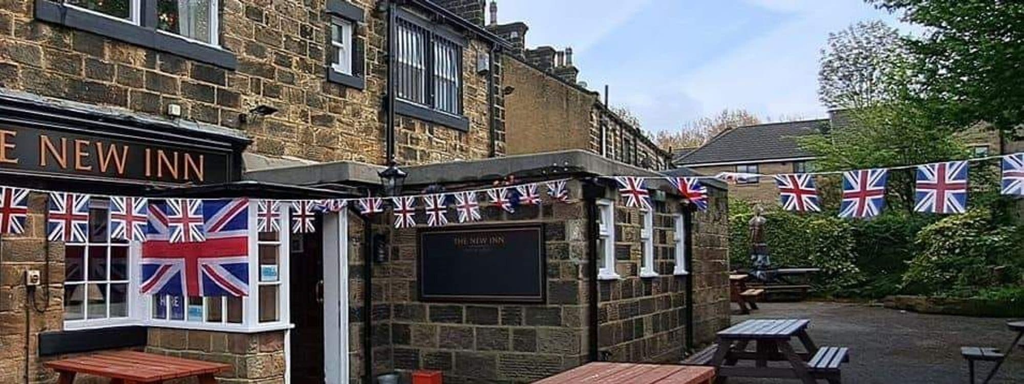Alt text: Exterior view of the New Inn – Yeadon, featuring a stone building adorned with Union Jack flags and outdoor seating. The scene includes a clear sky and greenery in the background, creating a welcoming atmosphere.