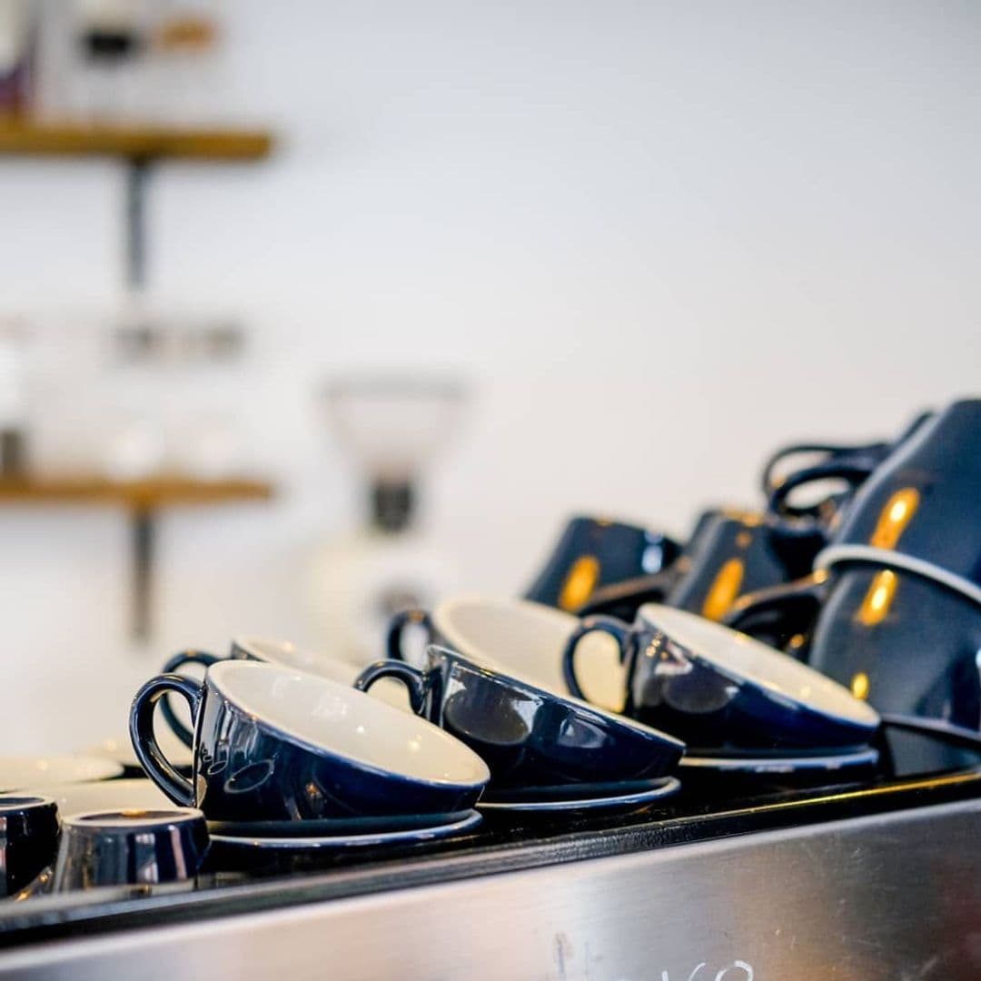 Stage Espresso Brewbar coffee cups barista cafe interior relaxation A stack of navy blue coffee cups and saucers is neatly arranged on a counter at Stage Espresso & Brewbar. The background features a simple, modern design with shelves holding various coffee-related items.