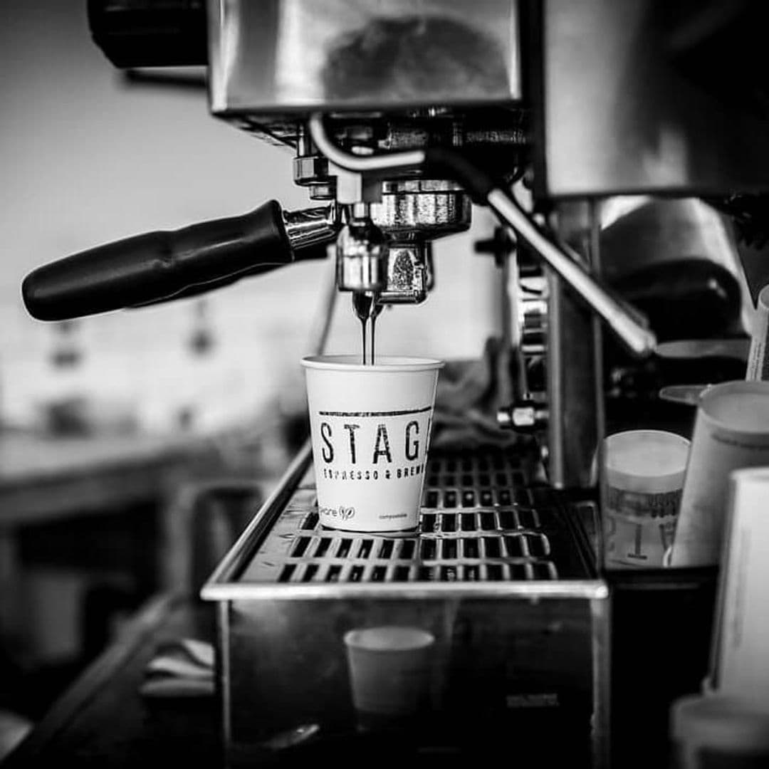 Stage Espresso Brewbar coffee machine brewed espresso cup black and white A close-up image of an espresso machine pouring coffee into a branded cup at Stage Espresso & Brewbar. The scene is in black and white, highlighting the coffee extraction process and the sleek design of the machine.