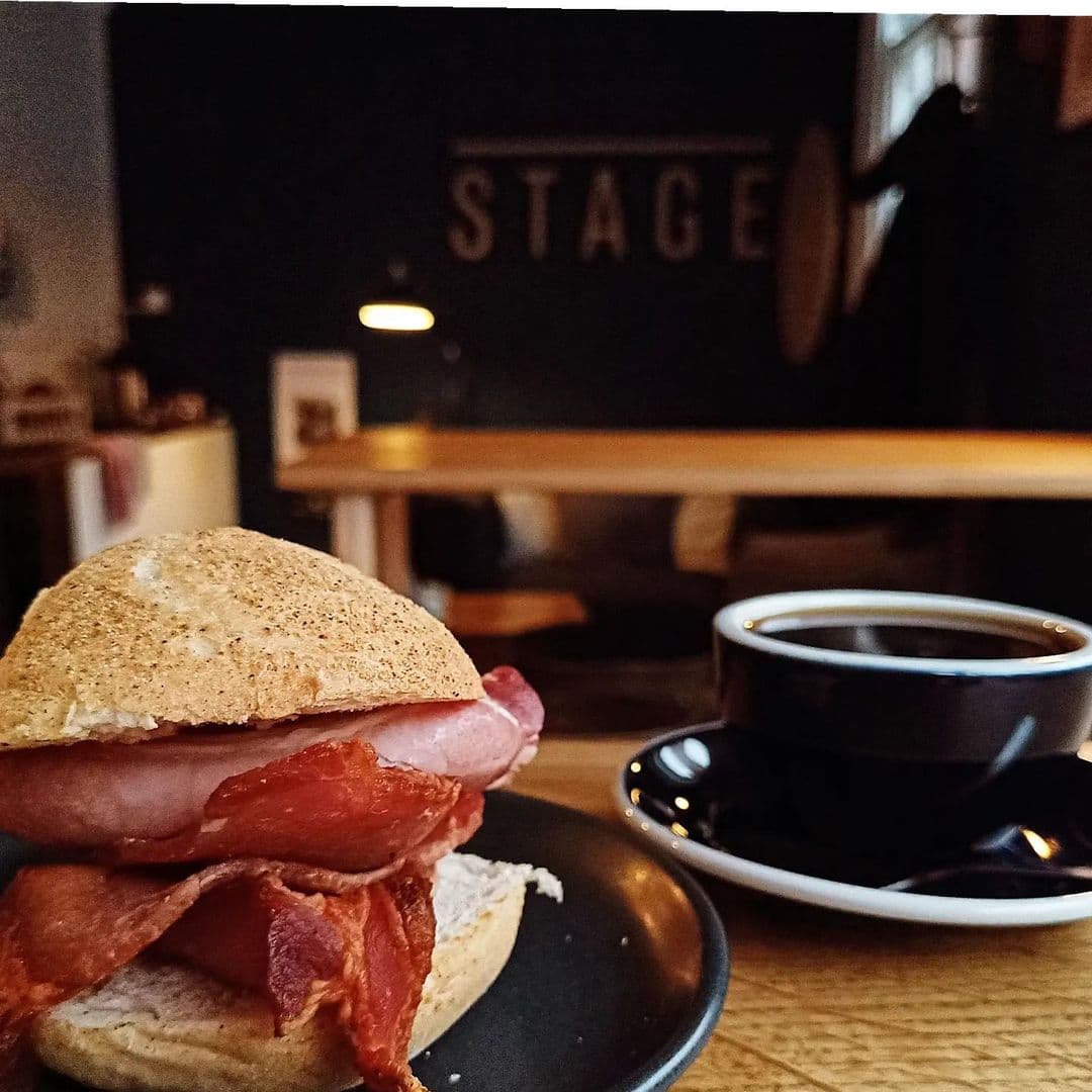 Stage Espresso Brewbar coffee sandwich breakfast cafe relaxation A close-up of a delicious sandwich with layers of bacon and ham on a whole grain bun, served on a black plate. In the background, a steaming cup of coffee sits on a wooden table, with a cozy café atmosphere featuring a dark wall that reads "STAGE." This image represents the inviting experience at Stage Espresso & Brewbar.