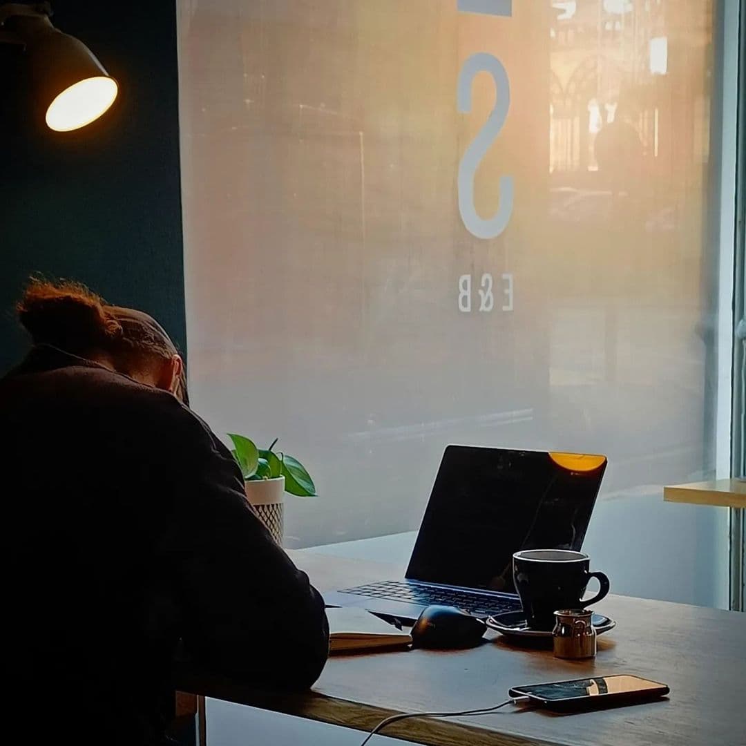 Stage Espresso Brewbar cozy workspace caffeine relaxation productivity A person sitting at a wooden table in Stage Espresso & Brewbar, focused on their work. A laptop is open in front of them, along with a notebook, a cup of coffee, and a small plant. Soft lighting creates a cozy atmosphere.
