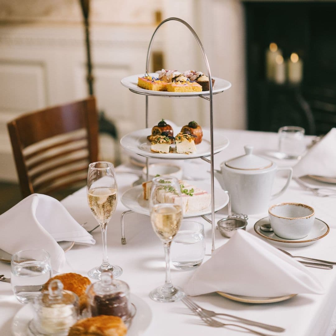 Terrace Tearoom afternoon tea desserts elegant setting A beautifully arranged afternoon tea setup at the Terrace Tearoom, featuring a tiered stand with assorted pastries, sandwiches, and scones. Two glasses of sparkling wine sit on the table alongside a teapot, cups, and neatly folded napkins, creating an inviting atmosphere for a delightful dining experience.