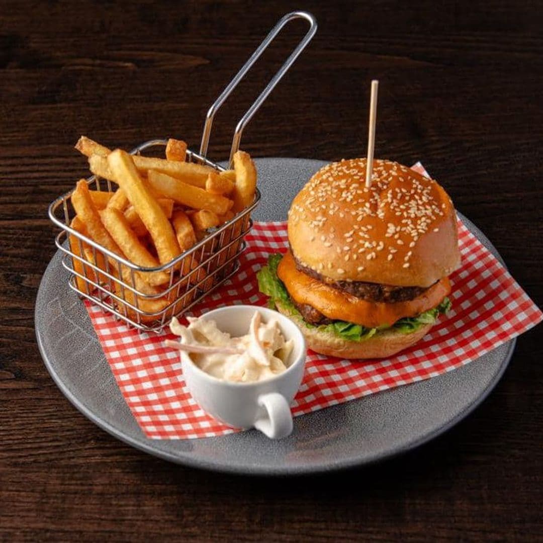 The Hare and Hounds Rothwell burger fries dining delicious food Delicious burger with cheese and lettuce, served on a sesame seed bun, accompanied by a side of crispy fries in a small basket and a cup of coleslaw. The presentation features a checkered paper liner on a round plate. The Hare and Hounds – Rothwell.