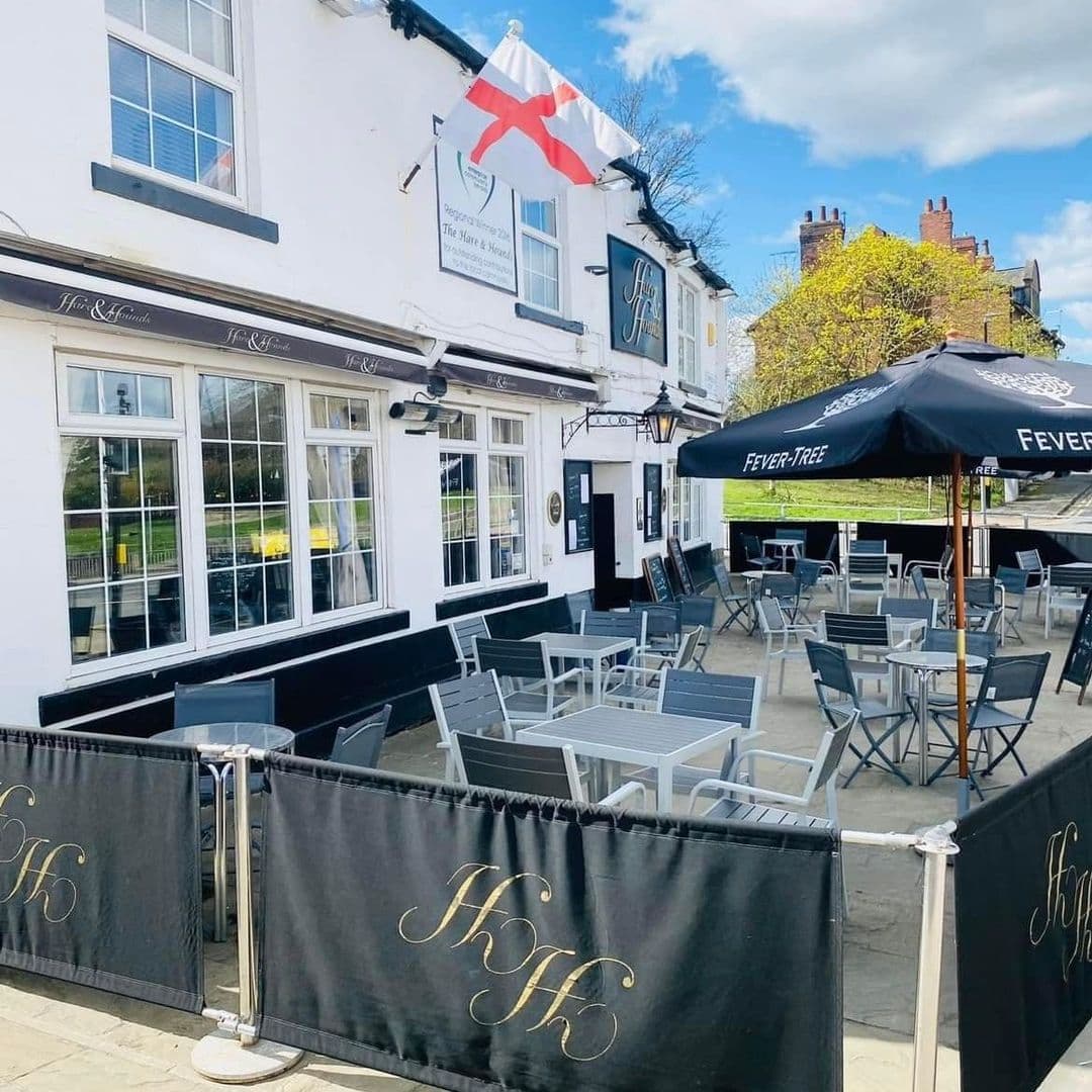 The Hare and Hounds Rothwell outdoor seating pub sunny day flag Alt text: Outdoor seating area at The Hare and Hounds – Rothwell, featuring tables and chairs under umbrellas, with a black barrier displaying the initials "HH" and an English flag flying above the pub.