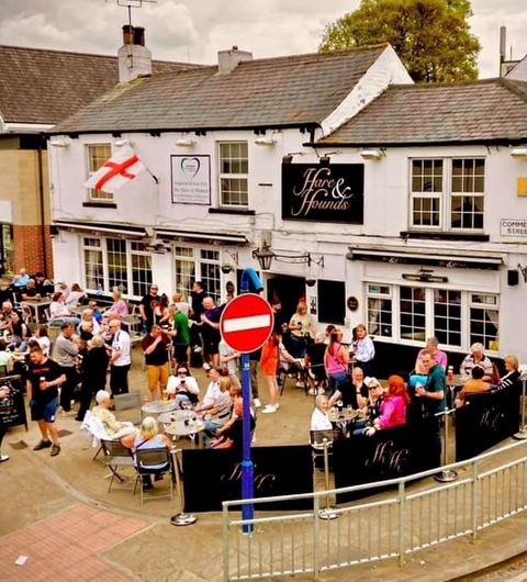 The Hare and Hounds Rothwell Alt text: A lively outdoor scene at The Hare and Hounds – Rothwell, featuring a crowd of people enjoying drinks and food at tables. The pub's exterior is visible, with the pub sign and an English flag displayed. A red no entry sign is in the foreground.