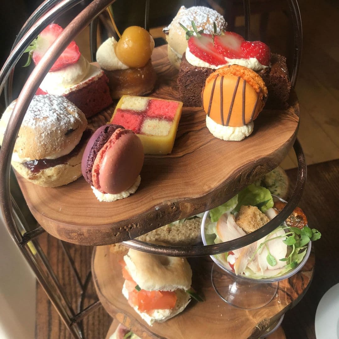 The Mansion afternoon tea desserts platter delicious treats A beautifully arranged tiered platter of assorted desserts and savory bites at The Mansion. The display includes scones with cream and jam, colorful macarons, small cakes, and a fresh salad, all presented on a wooden stand.