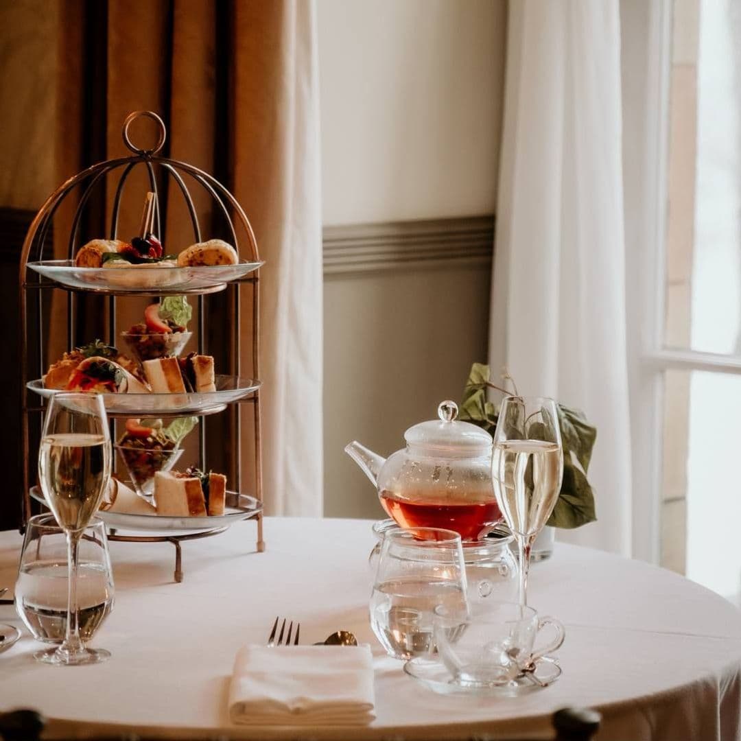 The Mansion afternoon tea elegant dining experience delicious food A beautifully set table at The Mansion featuring a tiered stand with an assortment of elegant finger foods, a teapot with steaming tea, and sparkling glasses of champagne. Soft natural light filters through a nearby window, creating a warm and inviting atmosphere.