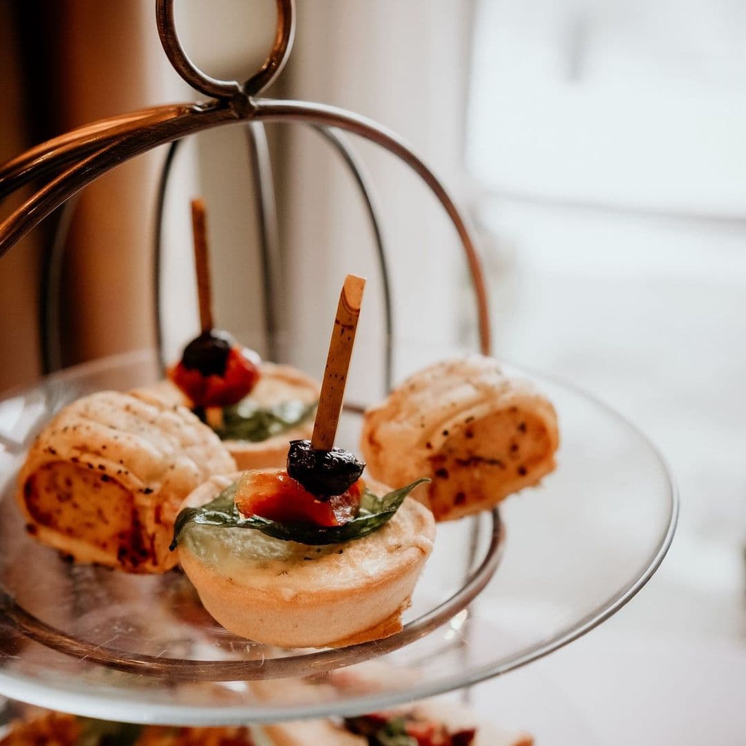 The Mansion gourmet appetizers elegant presentation tea time delight Delicious savory pastries served on a tiered stand, featuring a mix of ingredients like spinach, tomatoes, and olives. The setting is elegant, perfect for a dining experience at The Mansion.