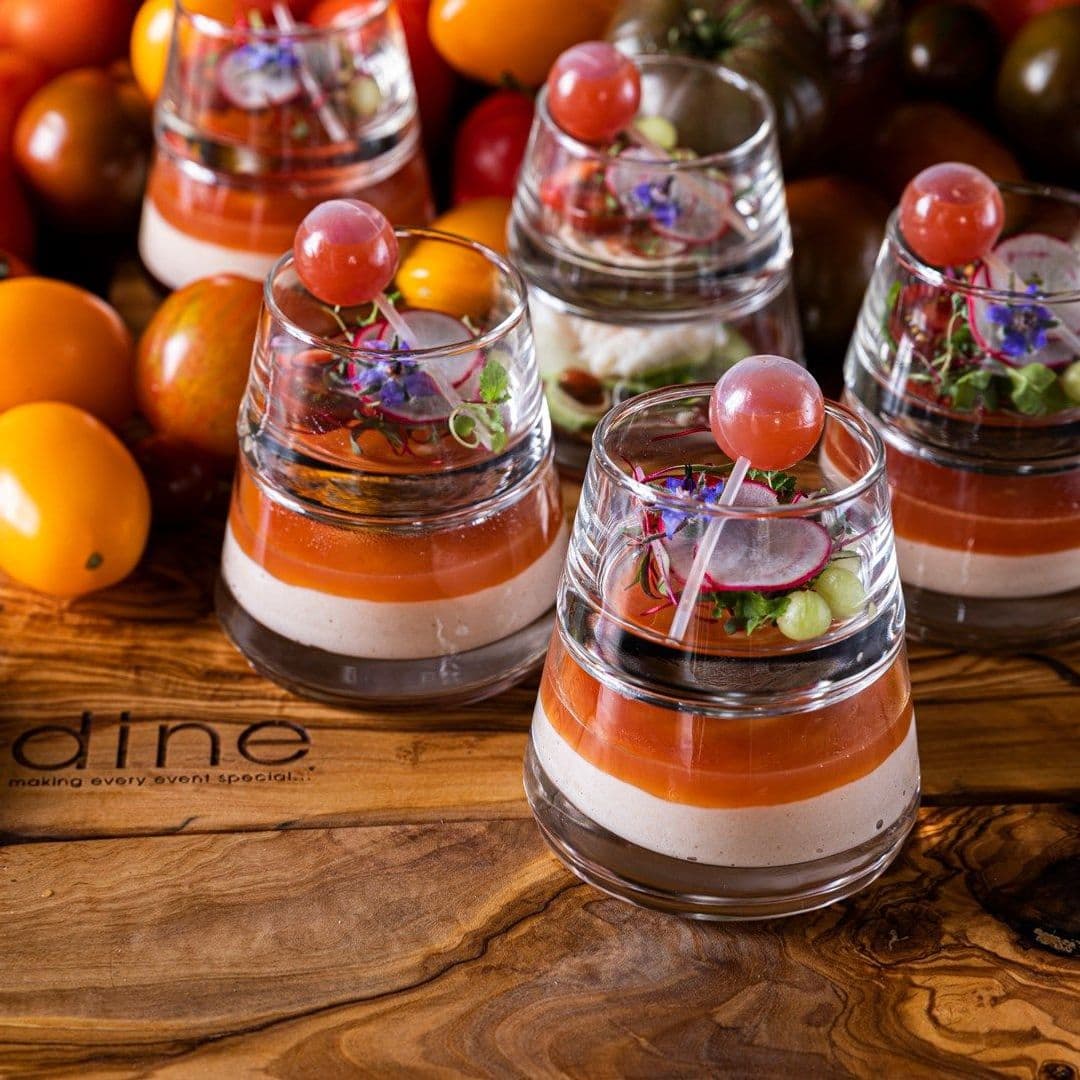 The Mansion gourmet dishes fresh ingredients culinary delight Delicious layered appetizers served in clear glasses, featuring vibrant colors and garnished with fresh herbs and vegetables. The background includes an assortment of colorful tomatoes. Perfect for a culinary experience at The Mansion.