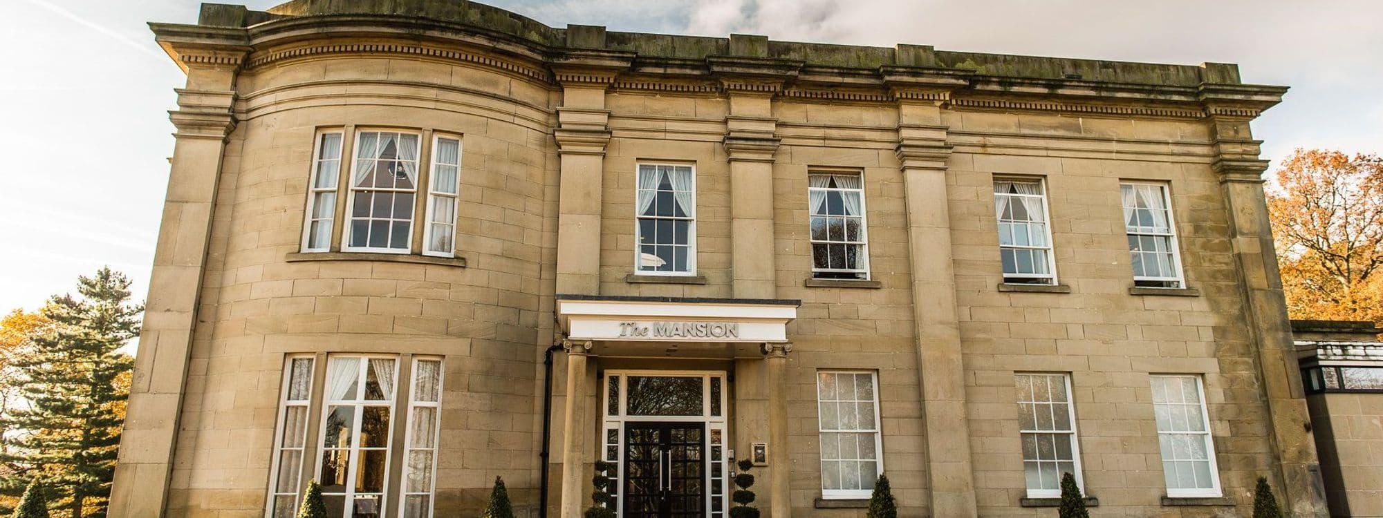 The Mansion, a beautiful historic building with elegant architecture, features large windows and a welcoming entrance. Surrounded by lush greenery, it showcases a blend of classic design and natural beauty, making it an ideal destination for visitors.