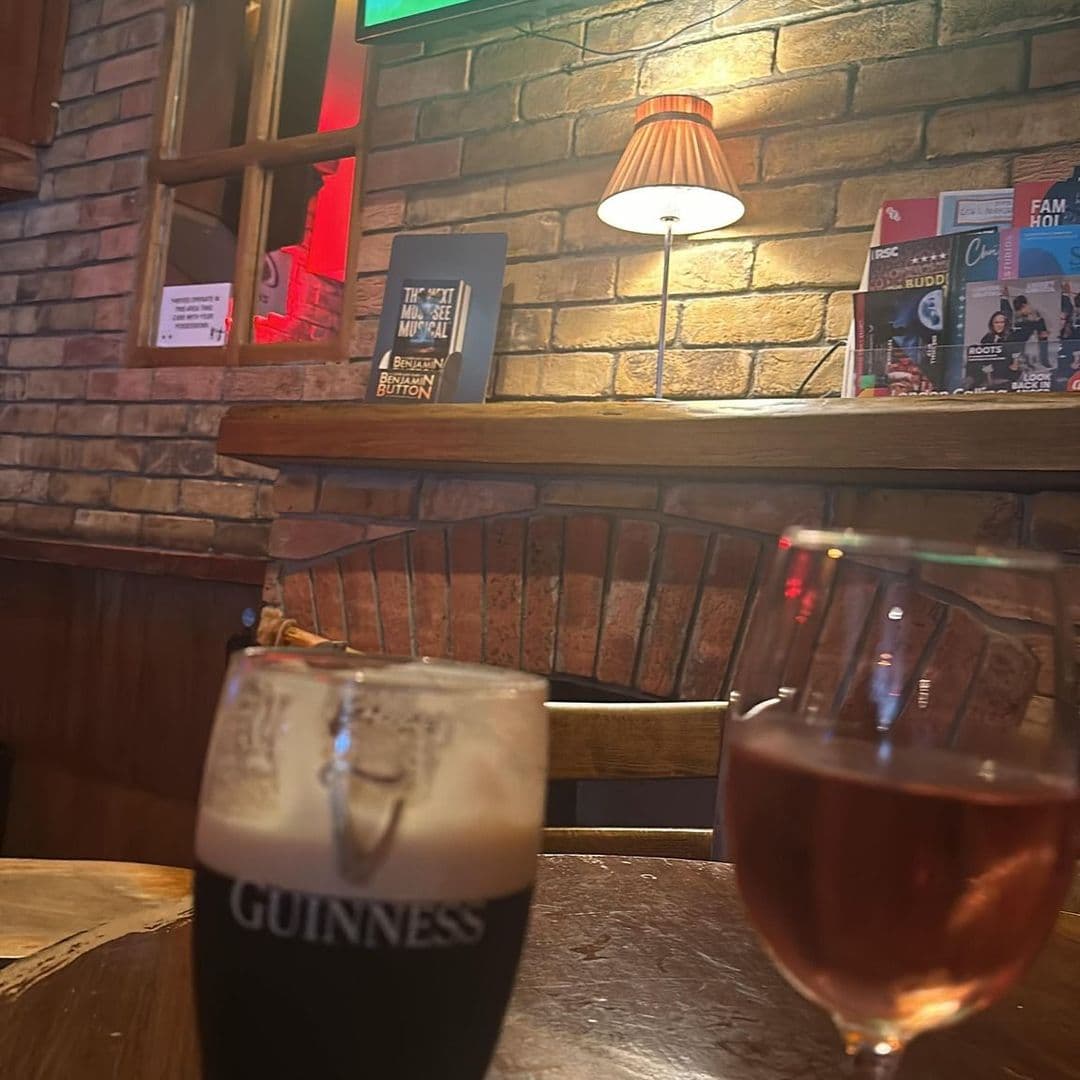 The Moot Hall Arms cozy bar experience drinks interior A cozy interior view of The Moot Hall Arms featuring a glass of Guinness and a glass of rosé wine on a wooden table. The warm ambiance is enhanced by a lamp and a brick wall, with a menu and promotional materials visible in the background.