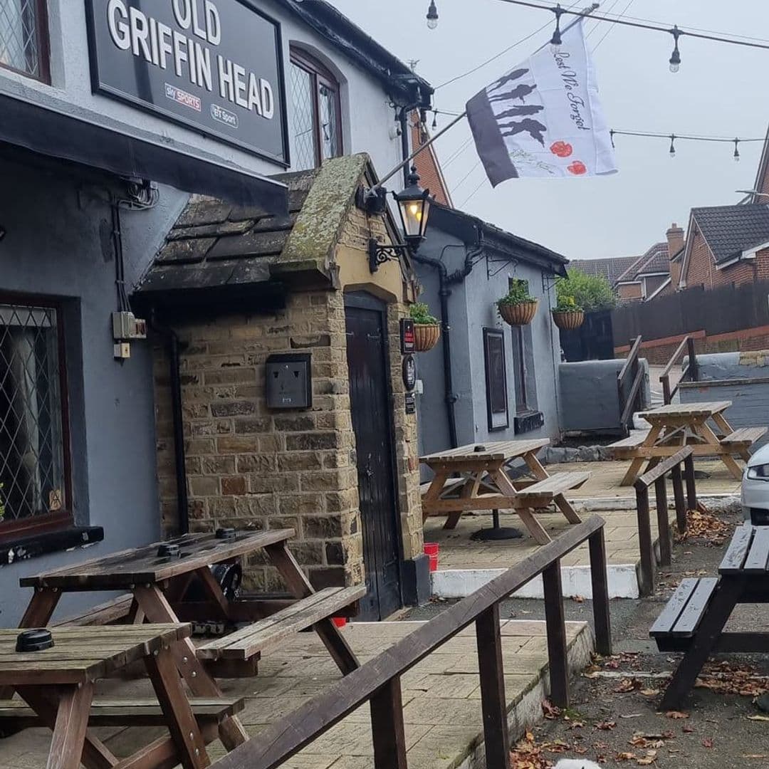 The Old Griffin Head pub exterior outdoor seating cozy atmosphere Alt text: The Old Griffin Head pub features a stone entrance, wooden picnic tables outside, and hanging flower pots. A flag is flying above the entrance, and the setting is calm with a few leaves on the ground.