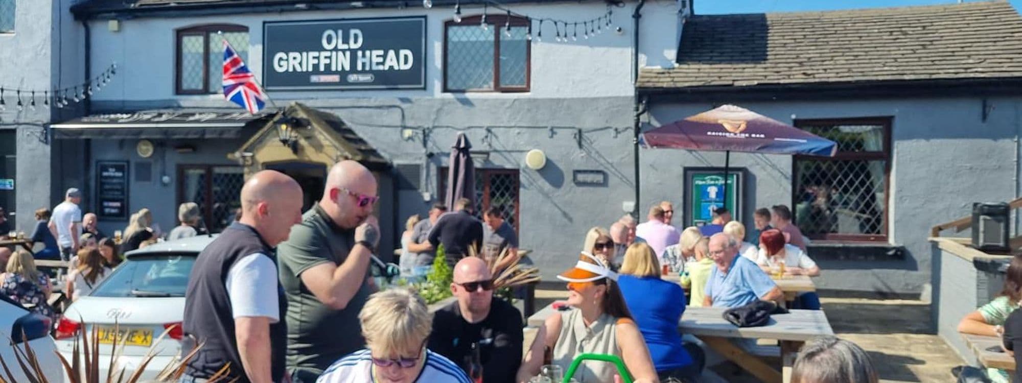 Alt text: A busy outdoor seating area at The Old Griffin Head pub, with people enjoying drinks and socializing under a sunny sky. The pub's sign is visible, along with a Union Jack flag.