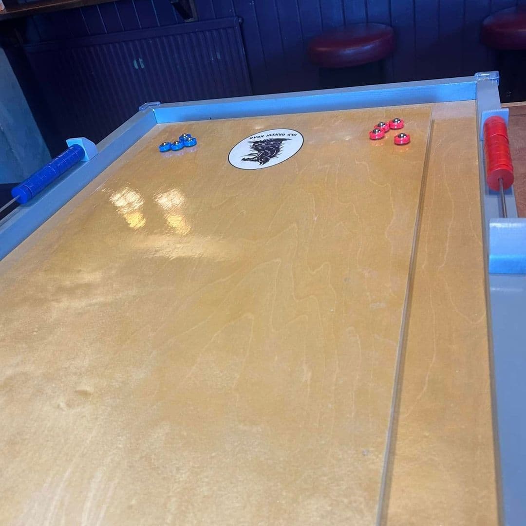 The Old Griffin Head shuffleboard game fun activity relaxation A close-up view of a wooden shuffleboard table at The Old Griffin Head, featuring blue and red pucks positioned on the surface. The table has a logo in the center and reflects soft lighting from the surroundings.