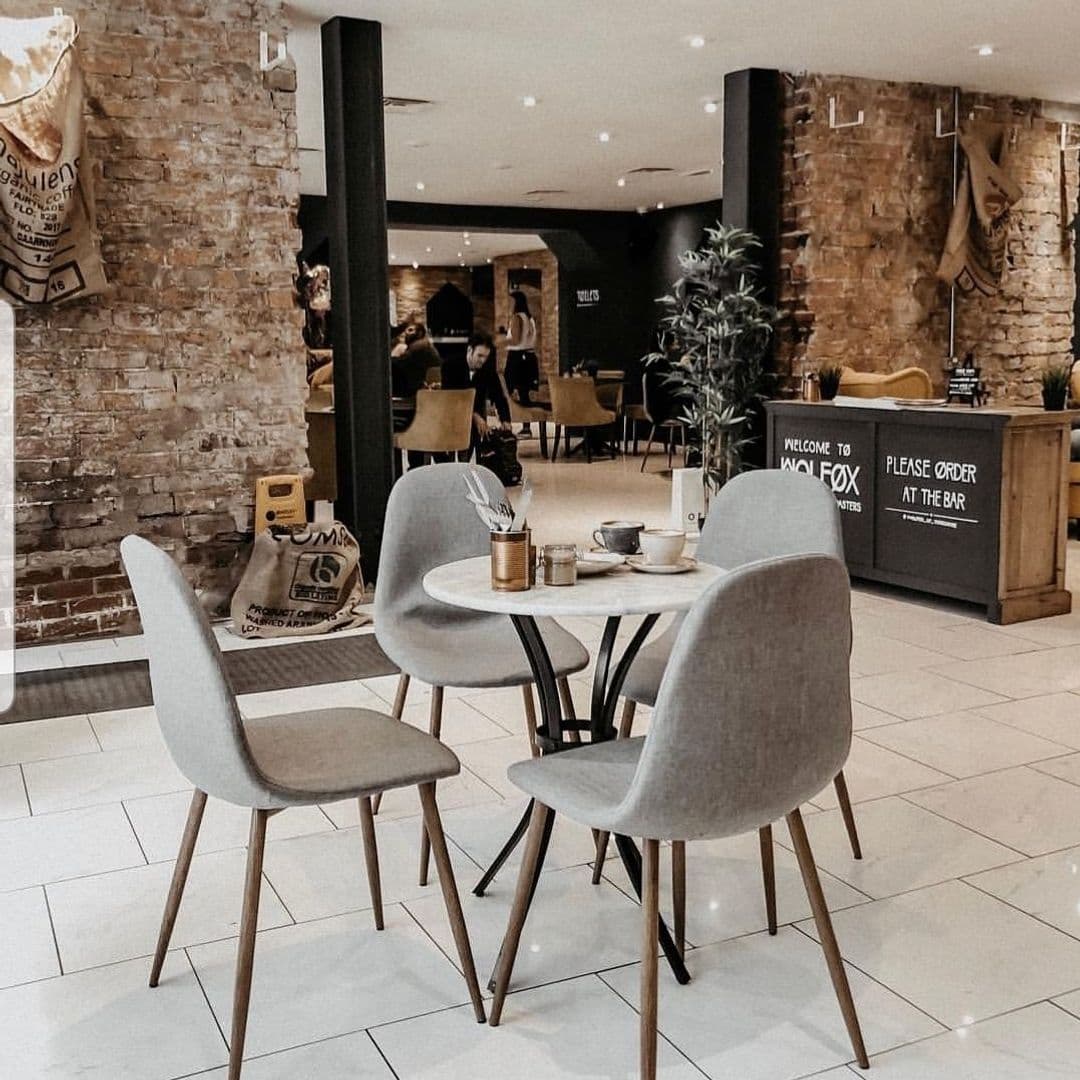 Wolfox Coffee cozy interior cafe modern design relaxing space Cozy interior of Wolfox Coffee featuring a small round table with two cups and a small plant. Surrounding are modern gray chairs and a rustic brick wall. The welcoming bar area is visible in the background with a sign that says "PLEASE ORDER AT THE BAR."