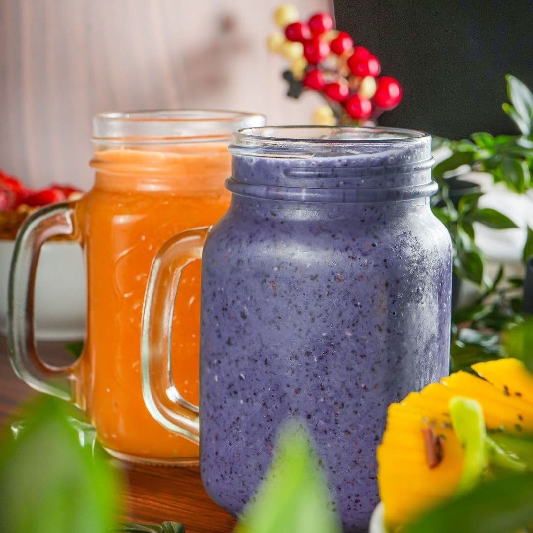 Wolfox Coffee fruit smoothies refreshing drinks vibrant colors Two colorful drinks in glass jars sit on a wooden table at Wolfox Coffee. One jar contains a bright orange beverage, while the other holds a rich purple smoothie. Fresh greenery and a slice of yellow fruit are visible in the foreground, adding to the vibrant atmosphere.