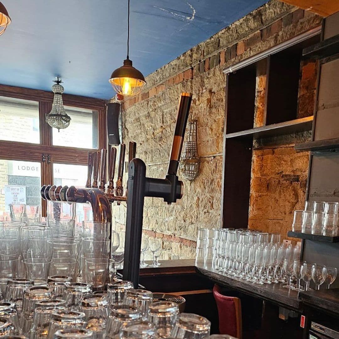 Beyond The Red Door bar interior glassware brewery ambiance Alt text: Interior view of a bar at Beyond The Red Door, featuring a row of beer taps, stacked glasses, and warm lighting against a rustic stone wall.