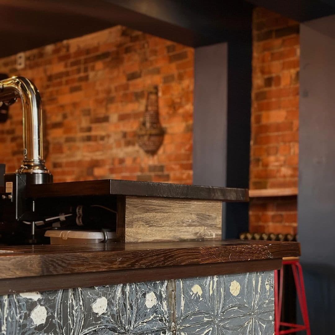 Beyond The Red Door cozy bar interior wooden counter brick wall modern Alt text: Interior view of a bar at Beyond The Red Door, featuring a wooden countertop and a backdrop of rustic brick walls. The bar has a polished look with decorative elements, creating a warm and inviting atmosphere.