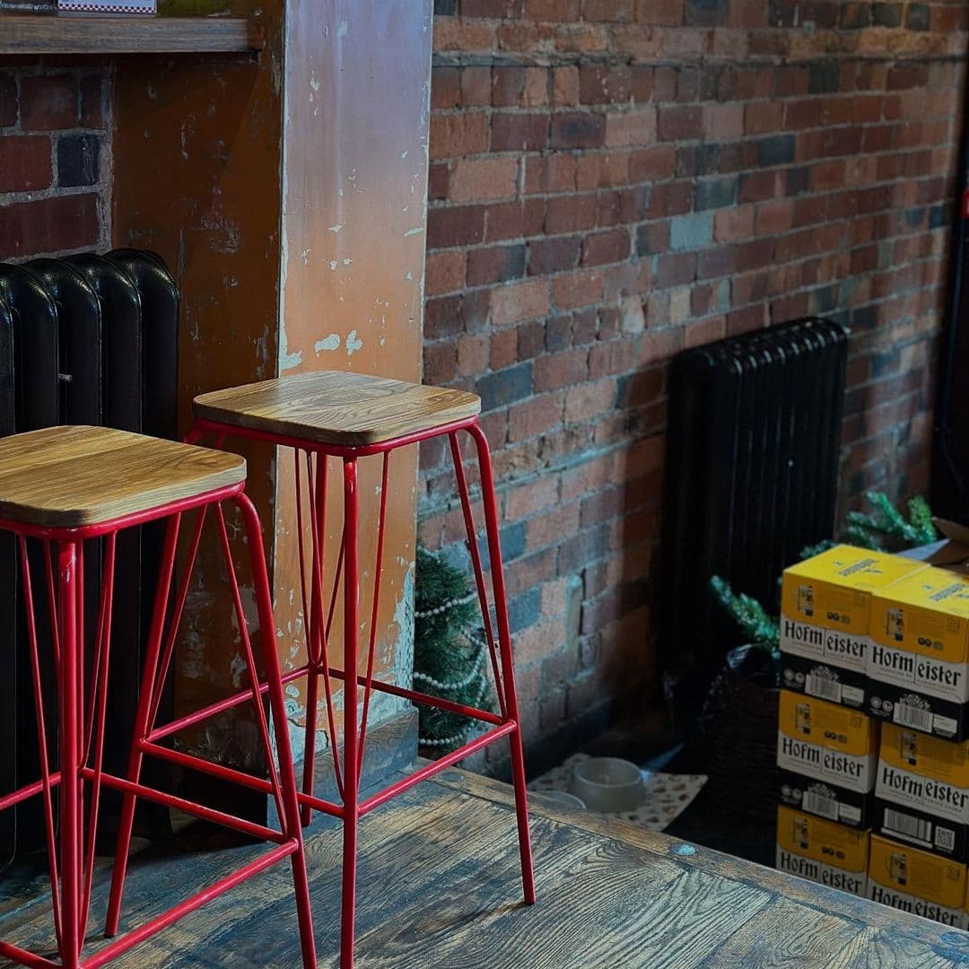 Beyond The Red Door cozy red bar stools interior design rustic ambiance Alt text: Two red metal bar stools with wooden seats positioned in front of a brick wall, alongside a vintage radiator and stacks of boxes, at Beyond The Red Door.
